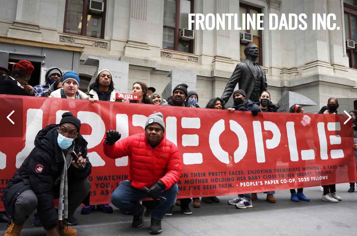 Diverse group of protesters outside a stone building holding a large red banner with white letters, some wearing masks and winter clothing.